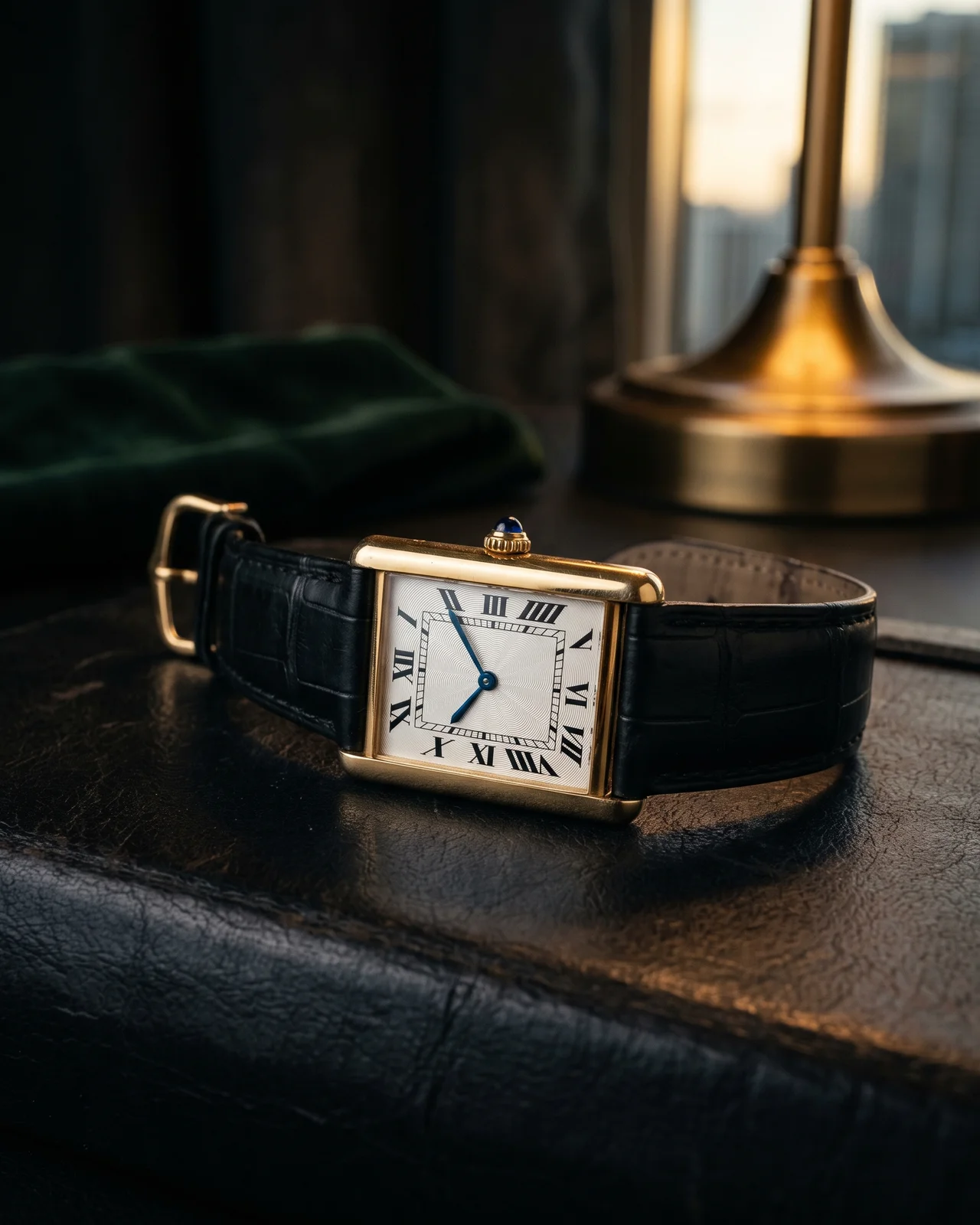 A rectangular Cartier Tank on a dark leather strap, lit from one side so the brancards and the blue cabochon crown stand out.