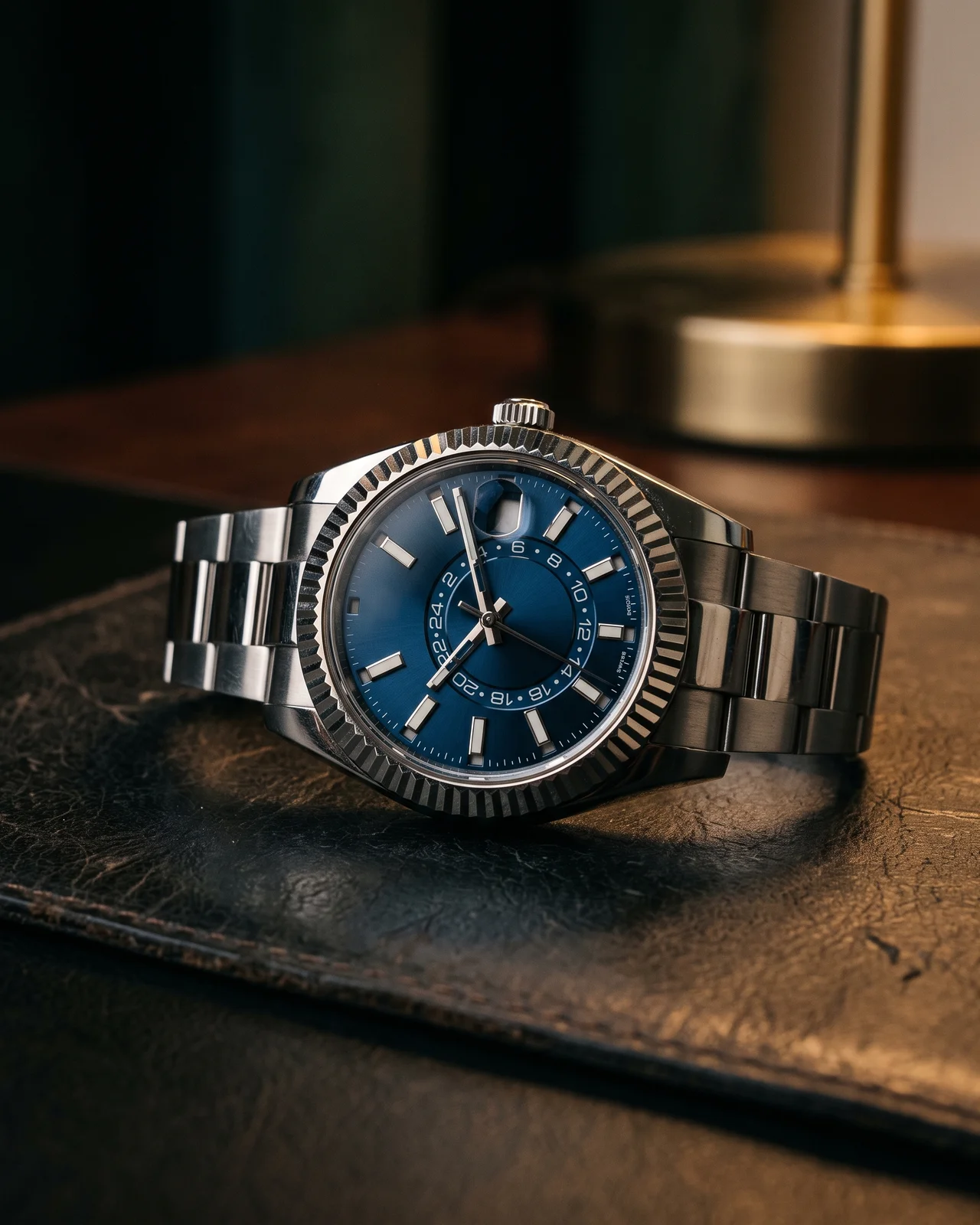 A steel Rolex Sky-Dweller with a white gold fluted bezel on a dark leather tray, the red month aperture and 24-hour disc visible.
